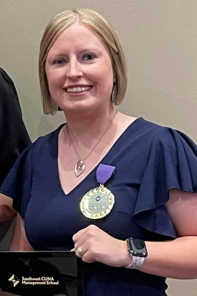 Person with chin length blond hair smiling as she holds a plaque and wears a large medal pinned to her blouse. 