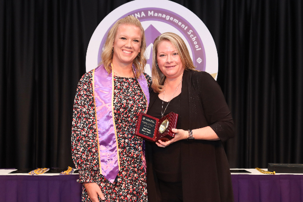 Two smiling people with shoulder length blond hair, one wearing a purple stole and the other holding a plaque